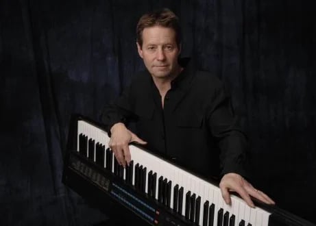 Man in black jacket playing a keyboard synthesizer against a dark background