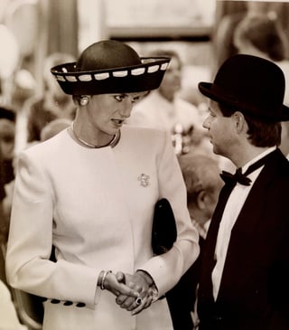 Black and white photo of two people in formal attire shaking hands at an event, woman in white dress and wide-brimmed hat, man in tuxedo with bowler hat